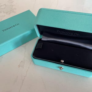 May include: An open, turquoise-colored box and matching rectangular box. The open box reveals a black interior with a silver clasp. The text "TIFFANY & Co." is printed on both boxes.