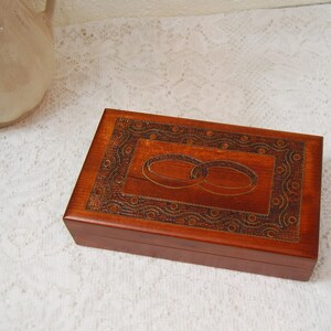 May include: A rectangular wooden box with a carved design featuring two interlocking rings. The box is a warm brown color and has a decorative border. The box is likely used for storing jewelry or small keepsakes.