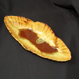 May include: A leaf-shaped ceramic dish with a glossy finish. The dish features a brown center, surrounded by a yellow and brown rim. A small sticker is visible on the dish. The dish is likely vintage.