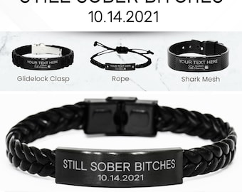 May include: Black leather bracelet with a silver clasp and a black metal plate engraved with the text "STILL SOBER BITCHES 10.14.2021". The bracelet is shown on a white marble background with the text "PERSONALIZED "SOBRIETY" BRACELET" below it.