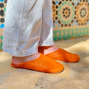 May include: A pair of bright orange leather slippers. The slippers are a slip-on style with a rounded toe. The person is wearing blue and white striped pants. The background features a decorative tile wall.