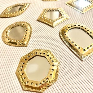 May include: A collection of small, decorative mirrors in various geometric shapes, including oval, diamond, and octagon. The mirrors have a gold-colored metal frame with a textured, hammered appearance. The mirrors are arranged on a light beige surface.