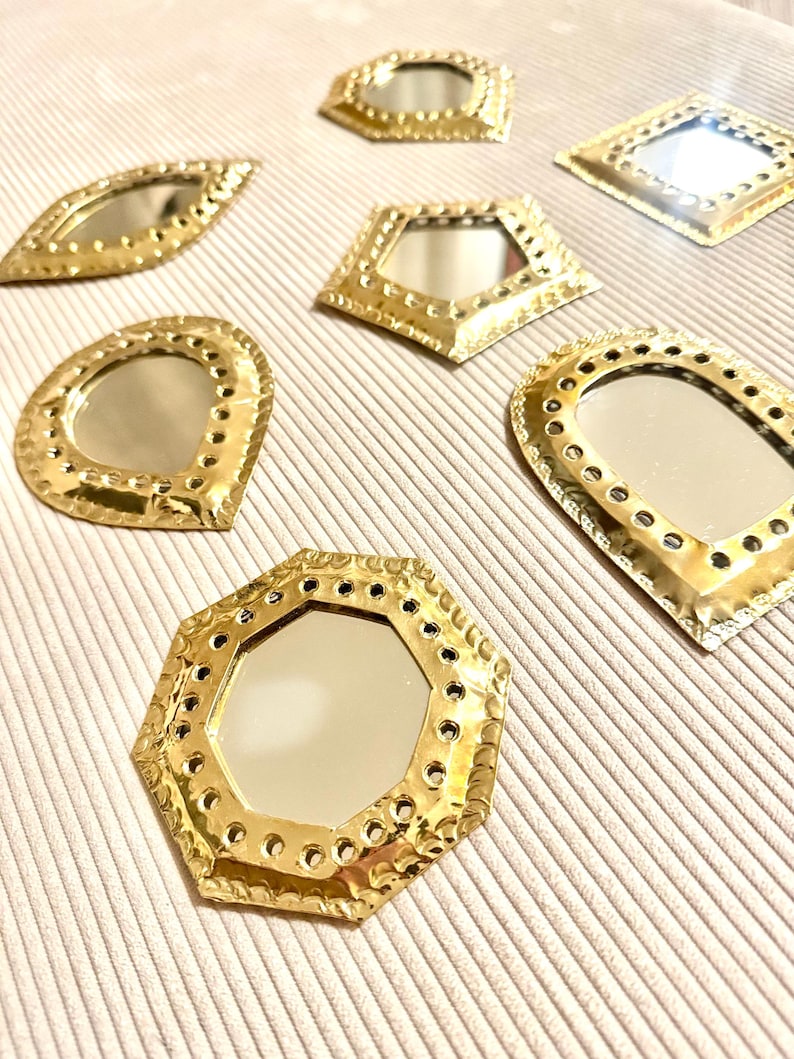 May include: A collection of small, decorative mirrors in various geometric shapes, including oval, diamond, and octagon. The mirrors have a gold-colored metal frame with a textured, hammered appearance. The mirrors are arranged on a light beige surface.