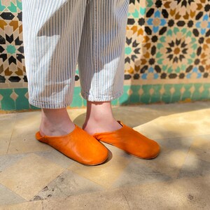 May include: A pair of bright orange leather slippers. The slippers are a vibrant, solid color and appear to be handmade. The person is wearing blue and white striped pants. The background features a decorative tile wall.