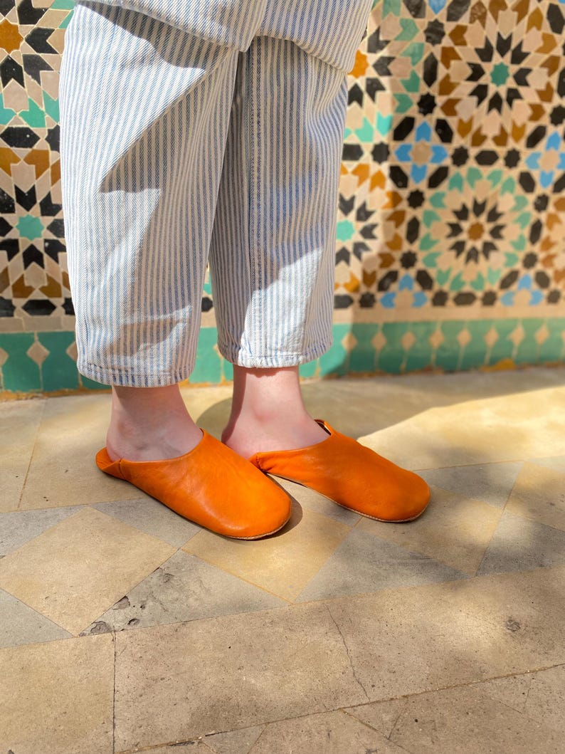 May include: A pair of bright orange leather slippers. The slippers are a vibrant, solid color and appear to be handmade. The person is wearing blue and white striped pants. The background features a decorative tile wall.