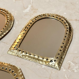 May include: Gold-colored, arched-top mirrors with a hammered metal frame and decorative edging. The mirrors have a reflective surface and are arranged on a marble-like surface. The frames feature small, evenly spaced holes.