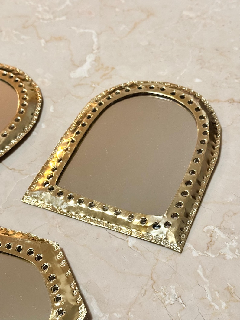 May include: Gold-colored, arched-top mirrors with a hammered metal frame and decorative edging. The mirrors have a reflective surface and are arranged on a marble-like surface. The frames feature small, evenly spaced holes.