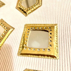 May include: Close-up of several small, gold-colored decorative mirrors in various geometric shapes, including square and octagonal. Each mirror has a reflective surface and a textured, dotted border. The mirrors are arranged on a light-colored, textured surface.