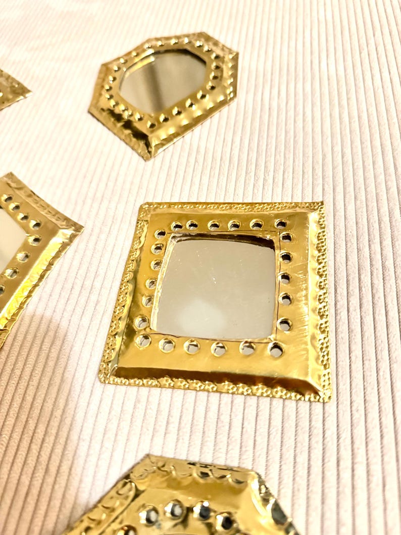 May include: Close-up of several small, gold-colored decorative mirrors in various geometric shapes, including square and octagonal. Each mirror has a reflective surface and a textured, dotted border. The mirrors are arranged on a light-colored, textured surface.