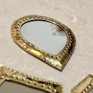 May include: A collection of decorative gold-colored mirrors with ornate, textured frames. The mirrors have a teardrop shape and are arranged on a light-colored surface. The frames feature a dotted pattern.