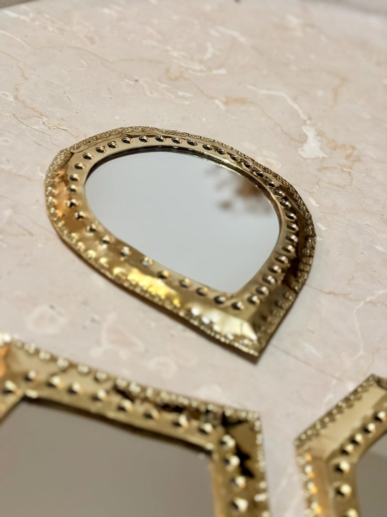 May include: A collection of decorative gold-colored mirrors with ornate, textured frames. The mirrors have a teardrop shape and are arranged on a light-colored surface. The frames feature a dotted pattern.