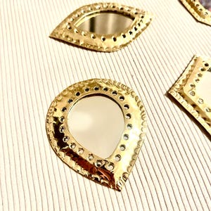 May include: Close-up of several gold-colored decorative mirrors in various shapes, including teardrop and eye shapes. Each mirror has a reflective surface and is surrounded by a textured, pierced metal frame. The mirrors are arranged on a striped, off-white surface.