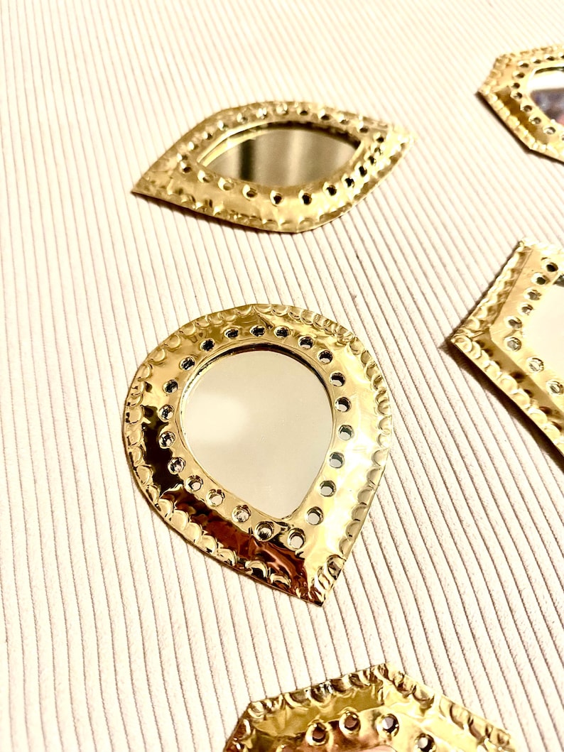 May include: Close-up of several gold-colored decorative mirrors in various shapes, including teardrop and eye shapes. Each mirror has a reflective surface and is surrounded by a textured, pierced metal frame. The mirrors are arranged on a striped, off-white surface.