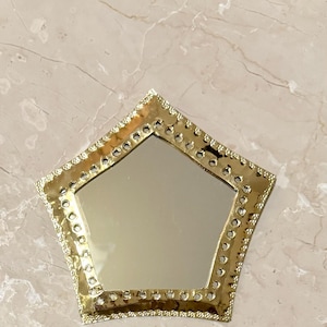 May include: A gold-colored, star-shaped mirror with a decorative, studded border. The mirror's reflective surface is surrounded by a detailed, ornate frame. The mirror is placed on a light-colored, marble-like surface.