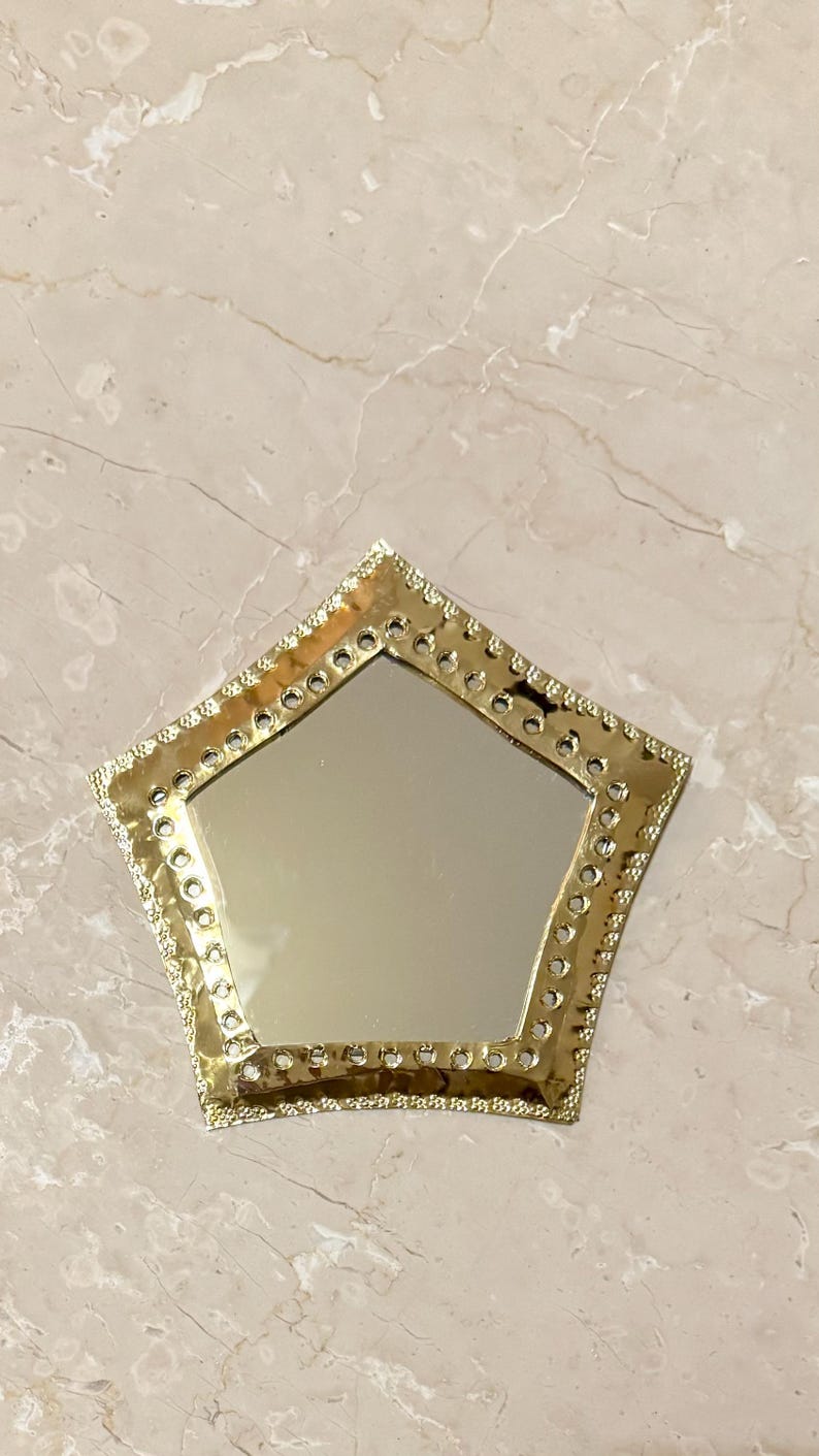 May include: A gold-colored, star-shaped mirror with a decorative, studded border. The mirror's reflective surface is surrounded by a detailed, ornate frame. The mirror is placed on a light-colored, marble-like surface.