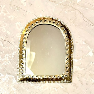 May include: A small, arched mirror with a gold-colored metal frame. The frame is decorated with small, circular accents around the edge. The mirror is set against a light-colored, marble-like surface.