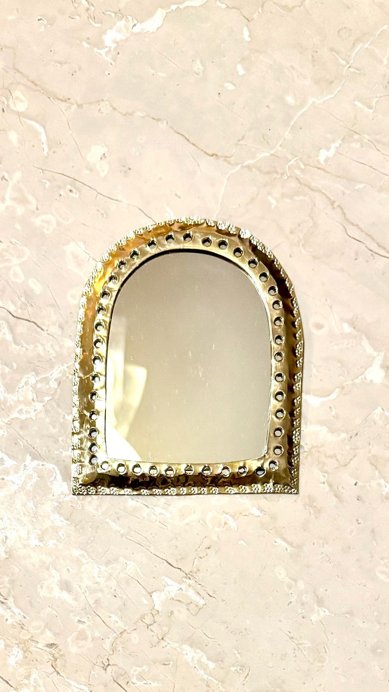 May include: A small, arched mirror with a gold-colored metal frame. The frame is decorated with small, circular accents around the edge. The mirror is set against a light-colored, marble-like surface.