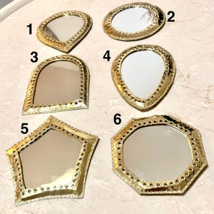 May include: A collection of six small, decorative mirrors with gold-colored frames. Each mirror has a unique shape, including oval, teardrop, and geometric designs. The frames feature a textured, dotted border, adding detail to the reflective surfaces.
