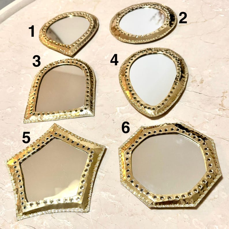 May include: A collection of six small, decorative mirrors with gold-colored frames. Each mirror has a unique shape, including oval, teardrop, and geometric designs. The frames feature a textured, dotted border, adding detail to the reflective surfaces.