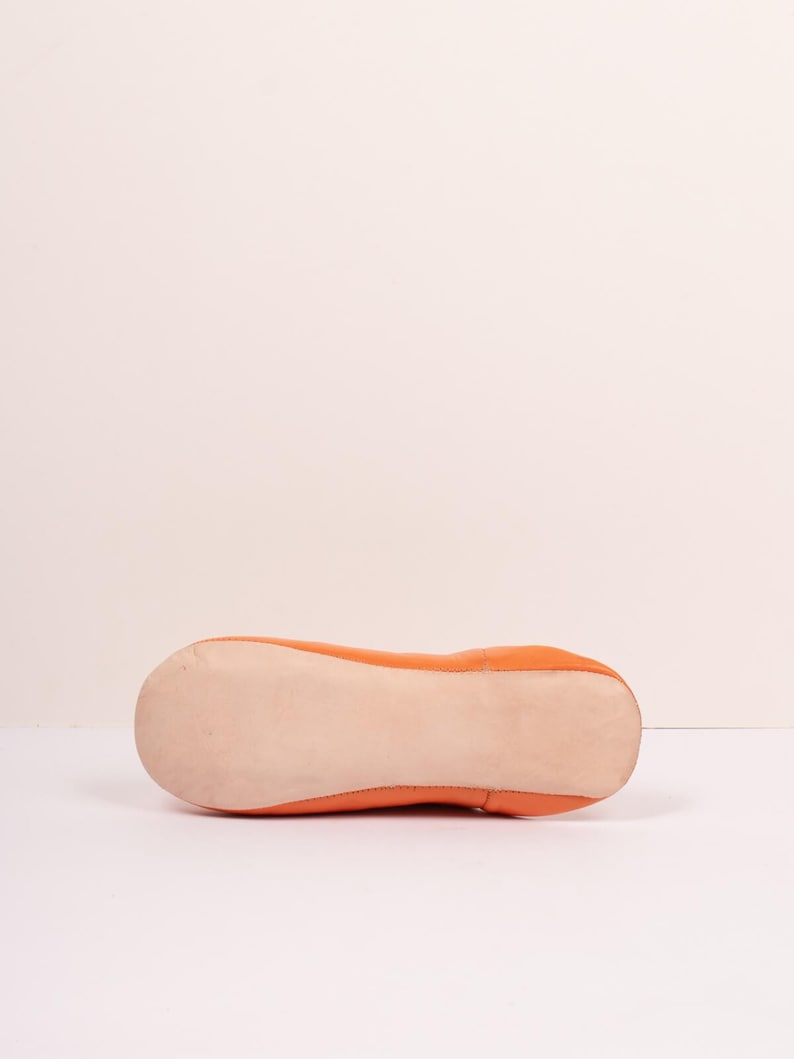 May include: Orange leather ballet flat. The shoe has a rounded toe and a flat sole. The sole is a light beige color, and the upper is a vibrant orange. The shoe is designed for comfort and style.