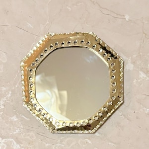May include: A gold-colored, octagonal mirror with a decorative, beaded edge. The mirror's frame has a series of small, circular openings. The mirror is set against a light-colored, marble-like surface.