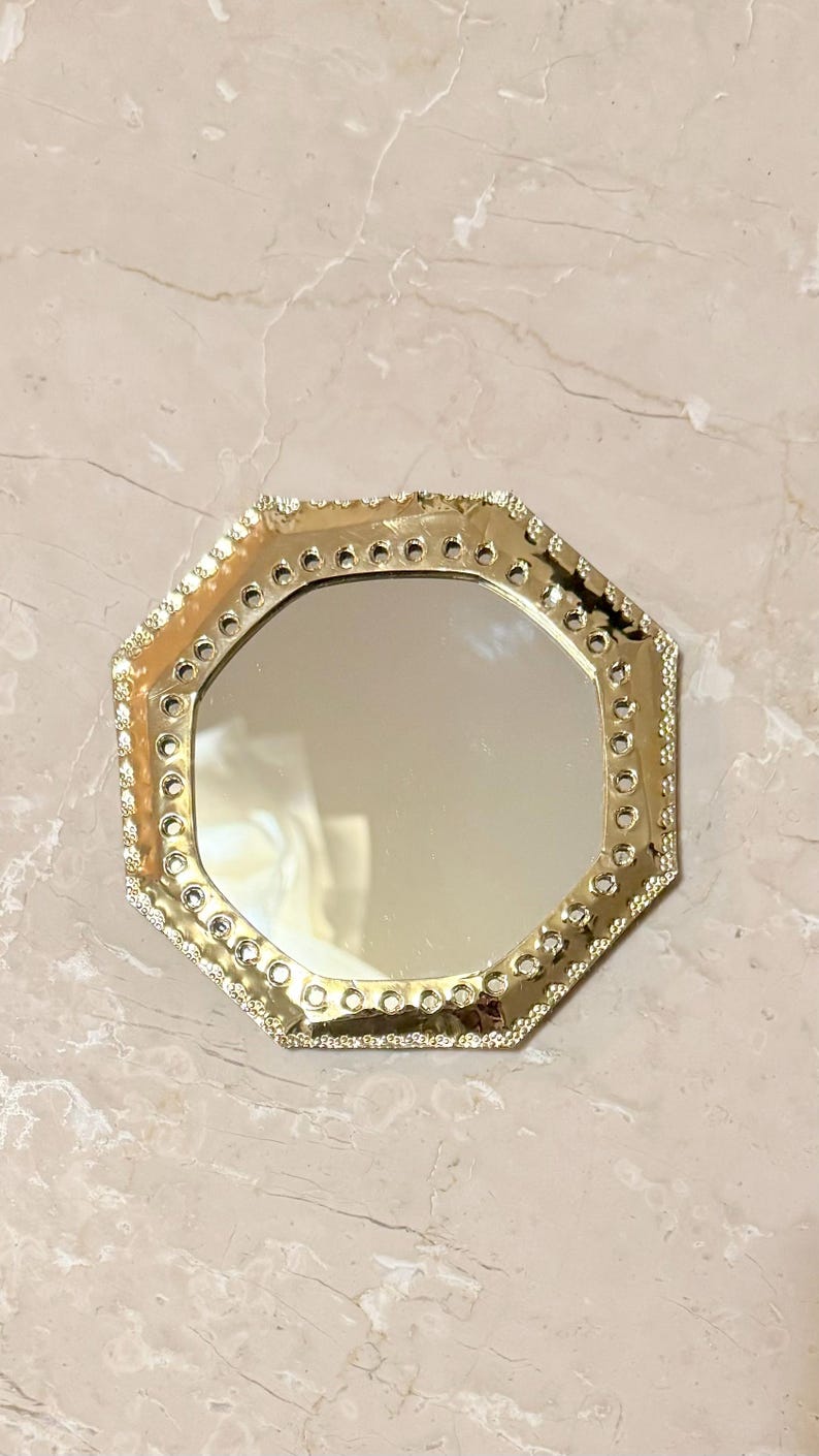 May include: A gold-colored, octagonal mirror with a decorative, beaded edge. The mirror's frame has a series of small, circular openings. The mirror is set against a light-colored, marble-like surface.