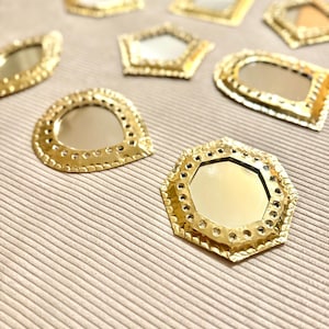 May include: A collection of small, gold-colored decorative mirrors in various geometric shapes, including a hexagon, a pentagon, and a rectangle. Each mirror has a reflective surface and is surrounded by a detailed, textured frame. The mirrors are arranged on a neutral-toned, textured surface.