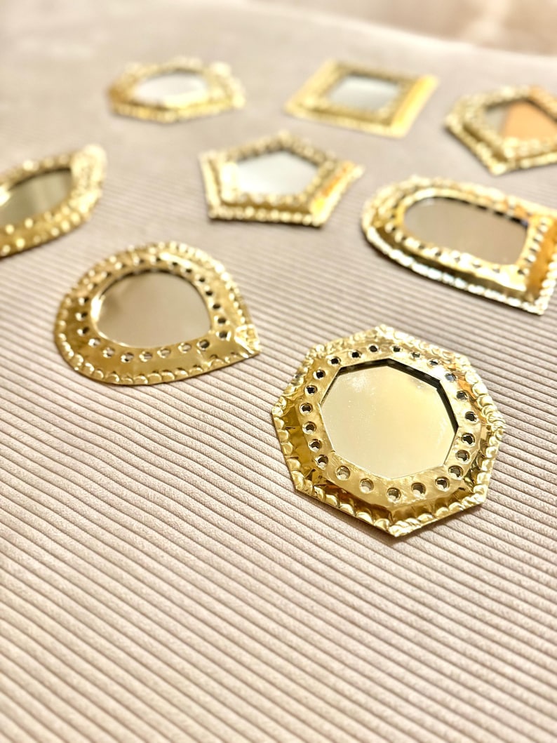 May include: A collection of small, gold-colored decorative mirrors in various geometric shapes, including a hexagon, a pentagon, and a rectangle. Each mirror has a reflective surface and is surrounded by a detailed, textured frame. The mirrors are arranged on a neutral-toned, textured surface.