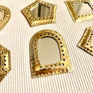 May include: A collection of small, decorative mirrors with gold-colored metal frames. The mirrors have various geometric shapes, including a pentagon, square, and arch. Each mirror has a reflective surface and is arranged on a textured, off-white surface.