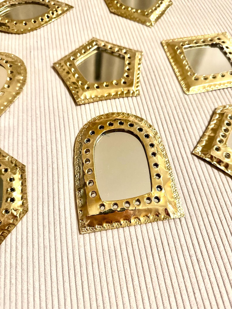 May include: A collection of small, decorative mirrors with gold-colored metal frames. The mirrors have various geometric shapes, including a pentagon, square, and arch. Each mirror has a reflective surface and is arranged on a textured, off-white surface.