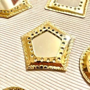 May include: Several gold-colored decorative mirrors in various shapes, including a pentagon, square, and circle. Each mirror has a reflective surface and a detailed, textured frame with small holes. The mirrors are arranged on a light beige, ribbed surface.