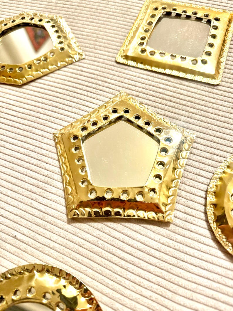 May include: Several gold-colored decorative mirrors in various shapes, including a pentagon, square, and circle. Each mirror has a reflective surface and a detailed, textured frame with small holes. The mirrors are arranged on a light beige, ribbed surface.