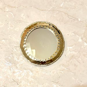 May include: A round, gold-colored compact mirror with a decorative, textured border. The mirror's edge is embellished with small, clear rhinestones. The mirror's surface is reflective, and the background is a marble-like pattern.