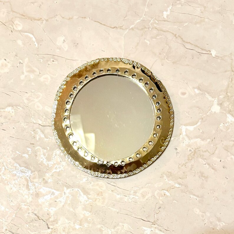 May include: A round, gold-colored compact mirror with a decorative, textured border. The mirror's edge is embellished with small, clear rhinestones. The mirror's surface is reflective, and the background is a marble-like pattern.
