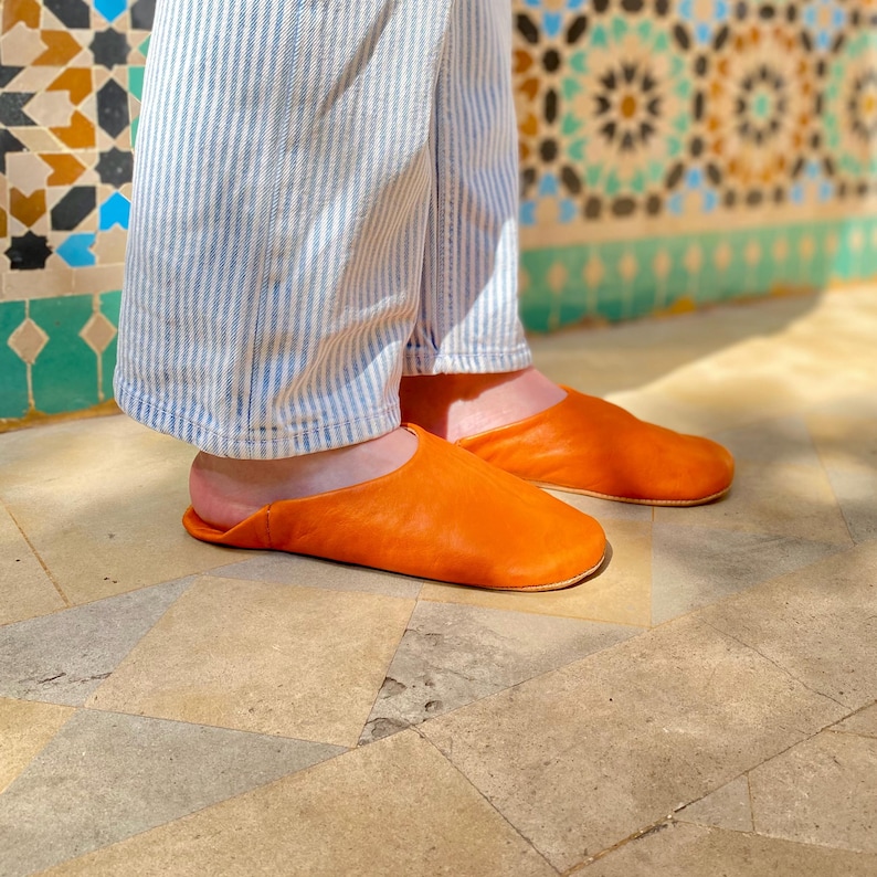 May include: A pair of bright orange leather slippers. The slippers are a slip-on style with a rounded toe. The person is wearing blue and white striped pants. The background features a decorative tile wall.