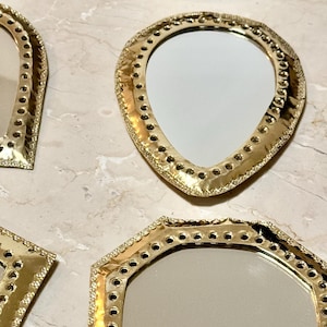 May include: A collection of decorative mirrors with gold-colored frames in various shapes, including round, teardrop, and geometric designs. The frames feature intricate detailing and small holes around the edges, reflecting light.