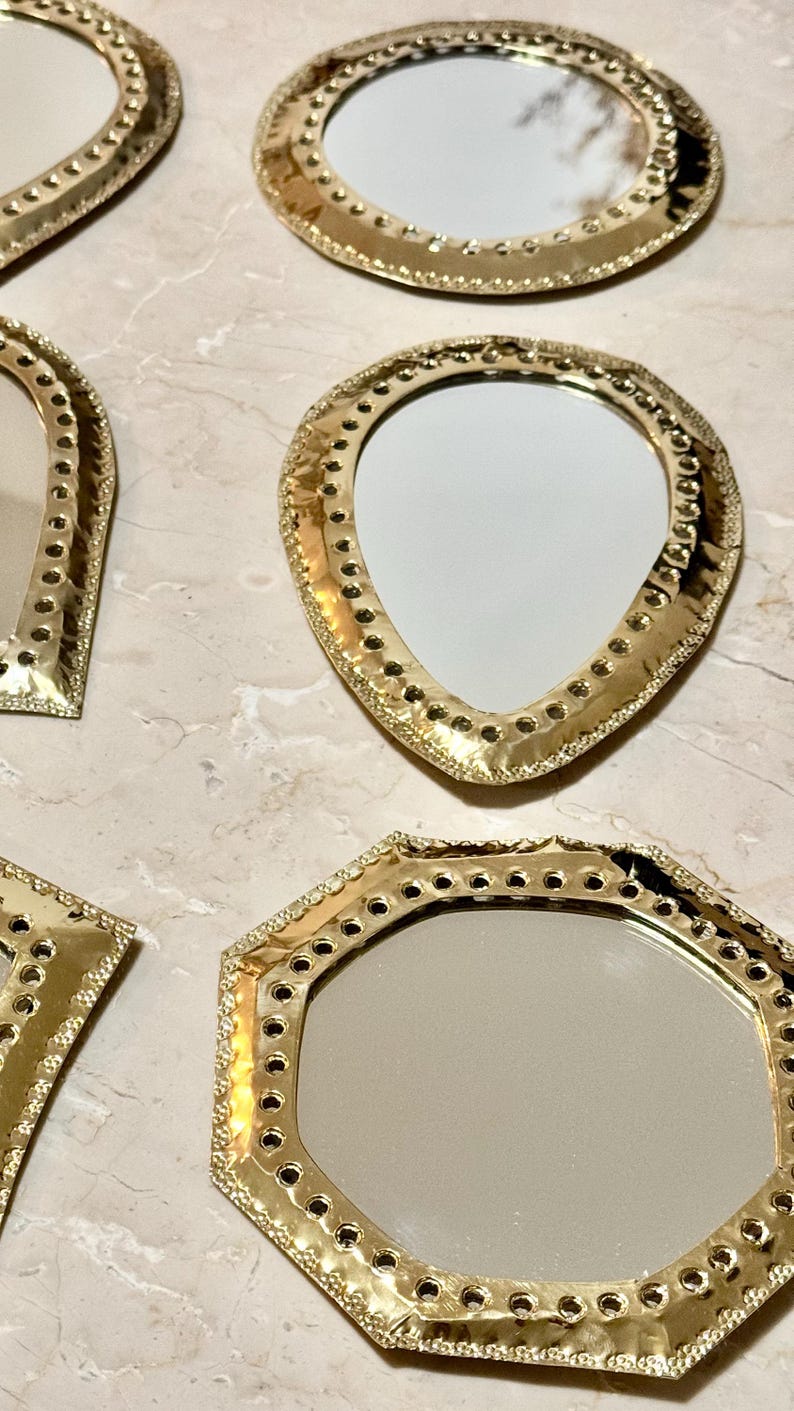 May include: A collection of decorative mirrors with gold-colored frames in various shapes, including round, teardrop, and geometric designs. The frames feature intricate detailing and small holes around the edges, reflecting light.