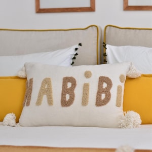 Op de afbeelding: Een wit kussen met het woord "HABIBI" geborduurd in bruin en beige garen. Het kussen heeft witte pompoenen op de hoeken.