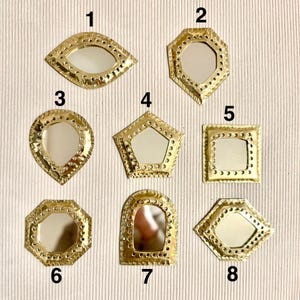 May include: A collection of eight small, decorative mirrors with gold-colored metal frames. The mirrors have various geometric shapes, including an eye, octagon, teardrop, pentagon, square, and arched shape. Each mirror has a reflective surface.