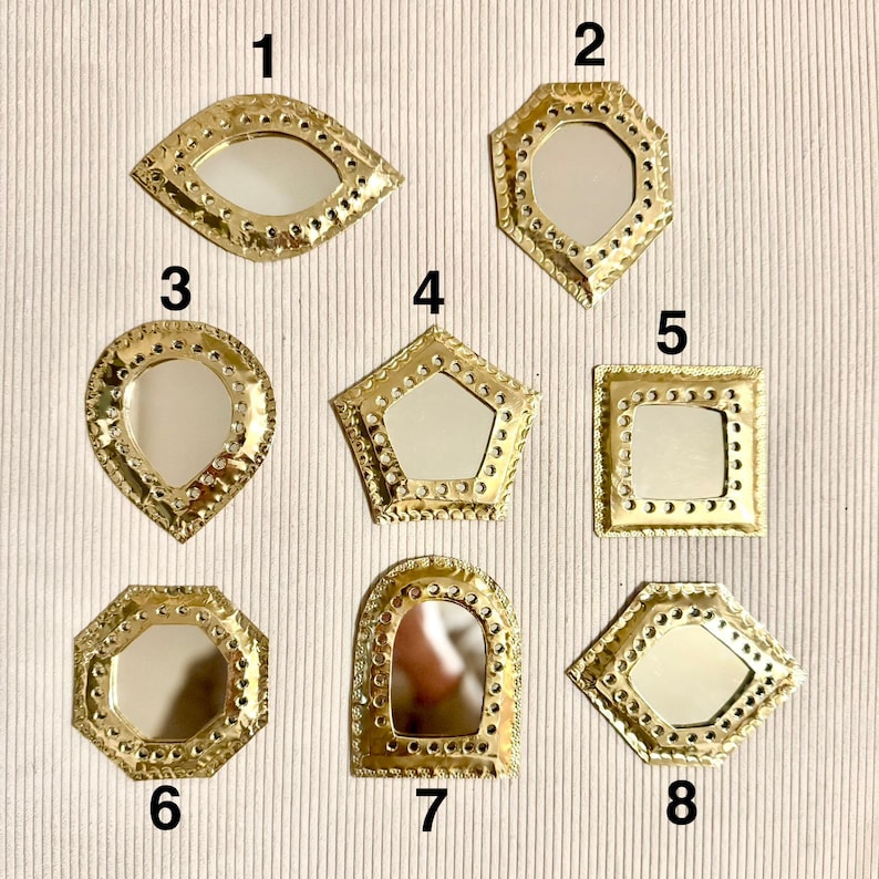May include: A collection of eight small, decorative mirrors with gold-colored metal frames. The mirrors have various geometric shapes, including an eye, octagon, teardrop, pentagon, square, and arched shape. Each mirror has a reflective surface.