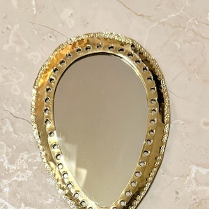 May include: A gold-colored, teardrop-shaped mirror with a decorative, textured border. The mirror's frame features a series of small, evenly spaced holes. The mirror is set against a marble-patterned surface.