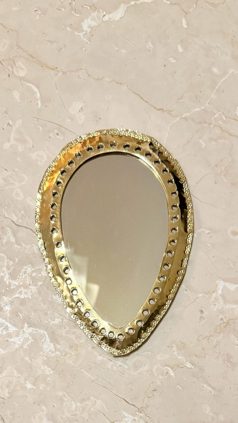 May include: A gold-colored, teardrop-shaped mirror with a decorative, textured border. The mirror's frame features a series of small, evenly spaced holes. The mirror is set against a marble-patterned surface.