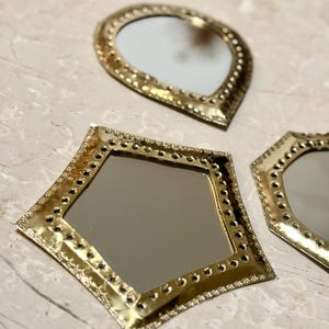 May include: Three decorative mirrors with gold-colored metal frames. The mirrors have different shapes: a teardrop, a diamond, and a hexagon. Each frame is detailed with small, evenly spaced holes around the edges.