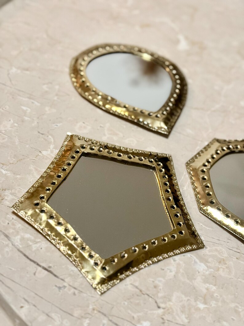 May include: Three decorative mirrors with gold-colored metal frames. The mirrors have different shapes: a teardrop, a diamond, and a hexagon. Each frame is detailed with small, evenly spaced holes around the edges.