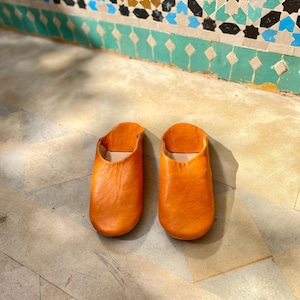 May include: A pair of orange leather slippers. The slippers are made of a soft, pliable leather and have a simple, slip-on design. The slippers are sitting on a stone floor with a decorative tile wall in the background.