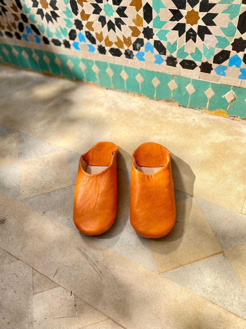May include: A pair of orange leather slippers. The slippers are made of a soft, pliable leather and have a simple, slip-on design. The slippers are sitting on a stone floor with a decorative tile wall in the background.