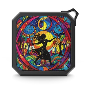 May include: A black, square-shaped portable speaker with a colorful stained-glass design. The design features a silhouette of a dancing figure, a crescent moon, and trees, with a rainbow of colors. The speaker has a black frame and a small loop for hanging.