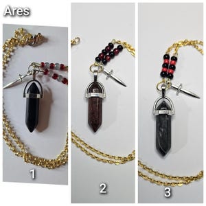 Puede incluir: Tres collares etiquetados como Ares, cada uno con cadena dorada, dije de espada plateado y colgante de cristal hexagonal. El collar 1 tiene un cristal negro y cuentas rojas, el 2 un cristal rojo oscuro y cuentas negras, y el 3 un cristal gris con cuentas.