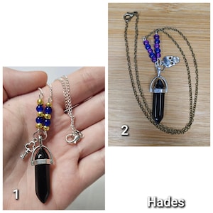 Puede incluir: Dos collares con colgantes de cristal negro en punta. El primero tiene una cadena plateada, cuentas azules y doradas y un dije de llave. El segundo tiene una cadena de bronce, cuentas moradas y azules y un dije de calavera, etiquetado Hades.