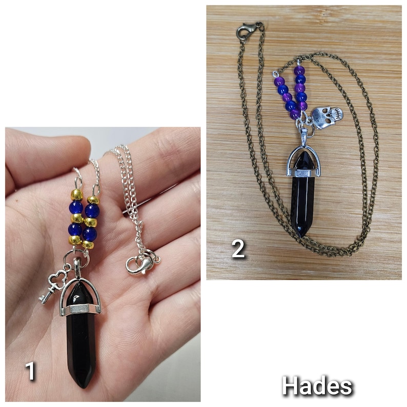 Puede incluir: Dos collares con colgantes de cristal negro en punta. El primero tiene una cadena plateada, cuentas azules y doradas y un dije de llave. El segundo tiene una cadena de bronce, cuentas moradas y azules y un dije de calavera, etiquetado Hades.